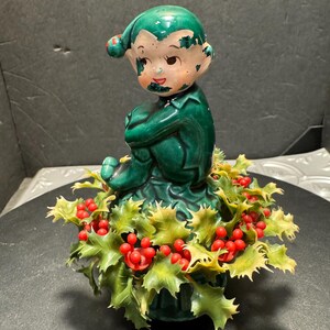 Op de afbeelding: Een vintage keramieken kerstelfbeeldje. De elf draagt een groene outfit en hoed met een rode pompon. De basis heeft groene hulstbladeren en rode bessen.
