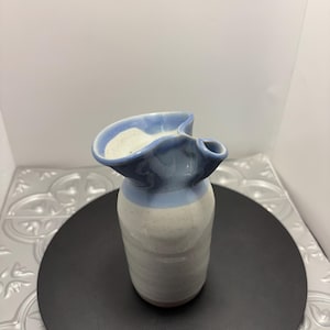 Puede incluir: Una pequeña jarra de cerámica hecha a mano con un borde ondulado único. La jarra presenta un diseño de dos tonos, con una parte superior azul claro y un cuerpo moteado blanco roto y gris. La jarra mide aproximadamente 13 cm de altura.