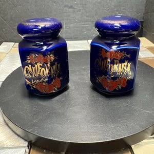 May include: Two cobalt blue ceramic jars with lids. Each jar has the word "CALIFORNIA" in gold lettering, with red floral accents. The jars are on a dark surface.