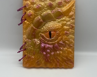 May include: A rectangular, fantasy-themed journal with a dragon eye design. The cover is a blend of orange, yellow, and pink hues, with a textured, scaly appearance. The eye is orange with a black slit pupil. The journal is bound with pink cord.