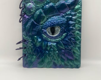 May include: A fantasy-themed journal with a dragon eye design. The cover is a mix of blue, green, and purple, with a textured, scaly appearance. The journal is bound with a dark purple cord.