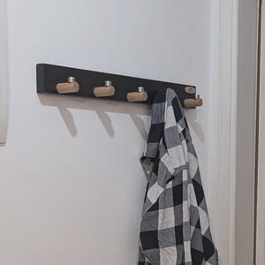 Puede incluir: Un perchero de pared negro con cuatro ganchos cilíndricos de madera y detalles metálicos en tono plateado. Una camisa de cuadros blancos y negros cuelga de uno de los ganchos, ocultando parcialmente la longitud y estructura del perchero.
