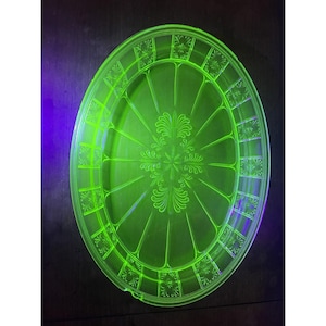 May include: A glowing green oval glass serving platter with a floral and geometric design. The platter has a central floral motif and a border of rectangular patterns. The glass appears to be vintage.