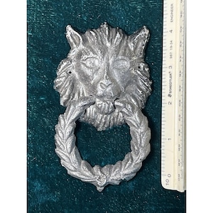 May include: A silver-colored door knocker in the shape of a lion's head with a ring-shaped handle. The knocker is made of metal and has a detailed design. The handle is a wreath. The knocker is approximately 4 inches in height.