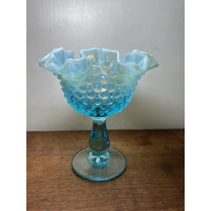 May include: A light blue opalescent glass compote dish featuring a hobnail texture on the bowl, a ruffled scalloped rim, and a pedestal base. The glass transitions from a translucent blue at the base to a milky, iridescent finish at the top.
