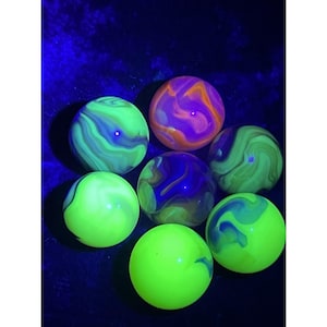 May include: Seven glowing marbles in shades of green, purple, and orange. The marbles have a swirled pattern and are set against a dark blue background. The marbles appear to be made of glass or a similar material.