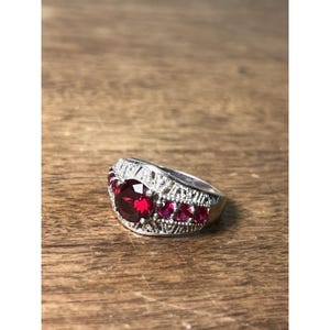May include: A silver ring with a large, oval, deep red gemstone at the center, flanked by smaller, round red stones. The band features intricate filigree detailing. The ring is set against a wooden surface.