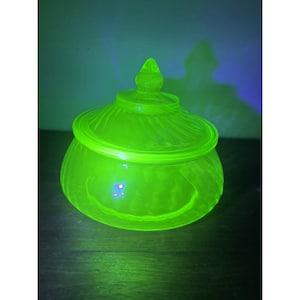 May include: A round, translucent glass bowl with a matching lid, glowing with a vibrant neon green color under ultraviolet light. The bowl and lid feature a subtle, swirling ribbed pattern and a pointed finial handle on the center of the lid.