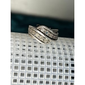 May include: A silver ring with a wave design, adorned with small, round, clear gemstones and a single blue gemstone. The ring is set on a white, grid-like surface, with a dark green background.