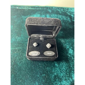 May include: Open black velvet jewelry box displaying a pair of square sterling silver stud earrings. The earrings are set with small, sparkling stones. The box has a label that reads "Sterling Silver".