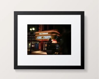 May include: A framed photograph of a building at night. The building has a marquee that reads "The Corner Food Court" in neon lights. A street lamp and a few trees are visible in the foreground. The image is framed in a black border.