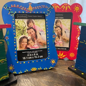 May include: Three colorful wooden picture frames with a whimsical folk art design. The frames are blue, red, and green with yellow, orange, and white accents. The blue frame has a photo of a family of four inside. The text on the blue frame reads "ArtMinds Frame I Cadre I Marco 4 in x 6 in 10.1 cm x 15.2 cm".