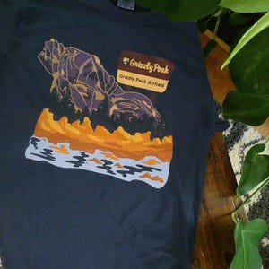 Puede incluir: Una camiseta azul oscuro con un gráfico de una cordillera y una puesta de sol. El gráfico incluye el texto "Grizzly Peak Airfield".