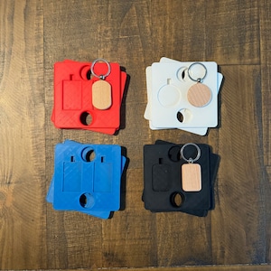 May include: Four different keychains with blank wooden tags. The keychains are displayed on a wooden surface. The keychains are red, white, blue, and black.