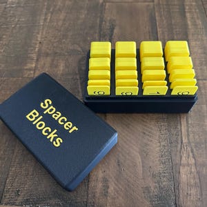 May include: A black case with the words "Spacer Blocks" in gold lettering sits next to a black tray filled with yellow spacer blocks. The blocks are rectangular and stacked. The background is a wooden surface.