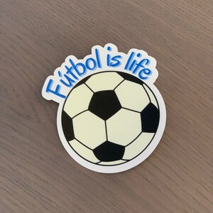 Pode incluir: Um autocolante com um design de bola de futebol com pent&aacute;gonos pretos e brancos. O texto "F&uacute;tbol is life" est&aacute; escrito em azul acima da bola. O autocolante est&aacute; numa superf&iacute;cie castanha.