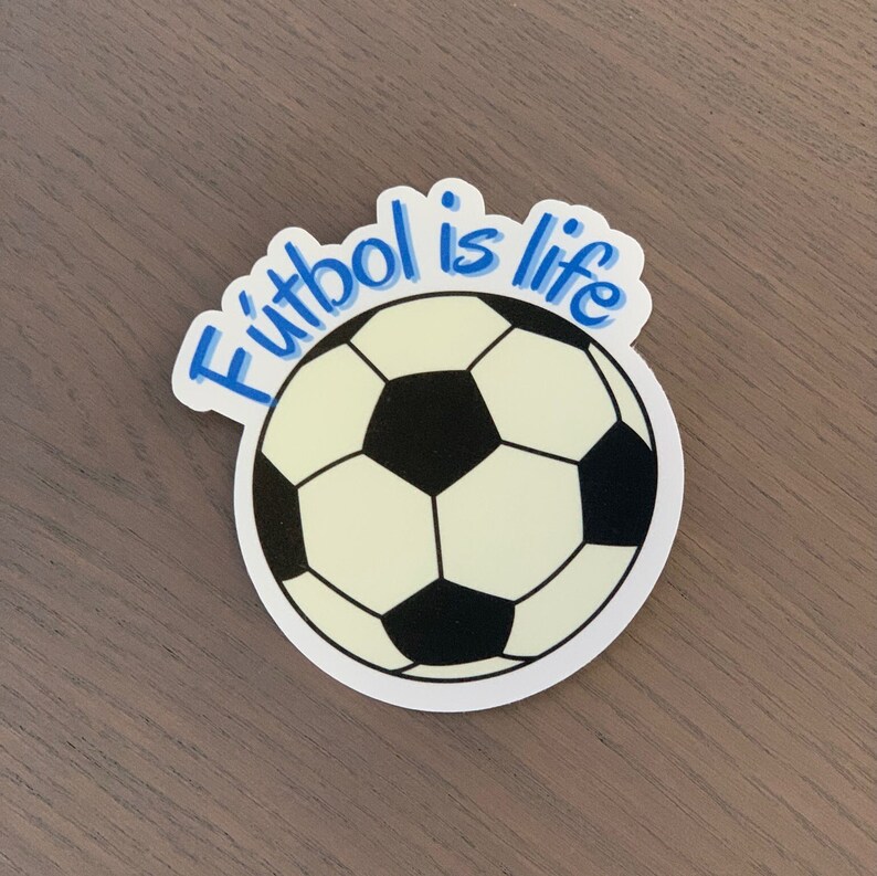Pode incluir: Um autocolante com um design de bola de futebol com pent&aacute;gonos pretos e brancos. O texto "F&uacute;tbol is life" est&aacute; escrito em azul acima da bola. O autocolante est&aacute; numa superf&iacute;cie castanha.