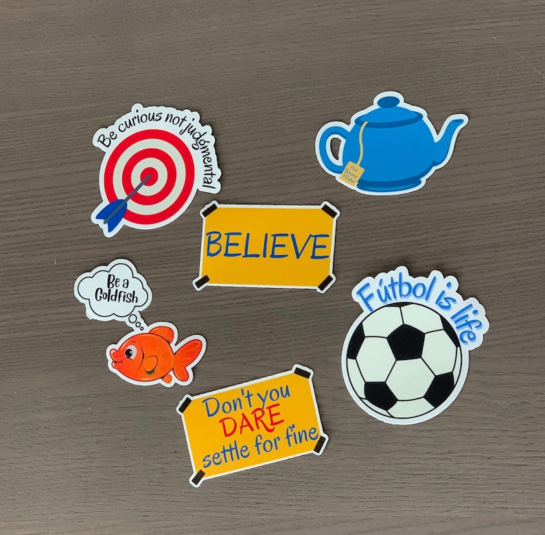 Pode incluir: Uma cole&ccedil;&atilde;o de autocolantes coloridos com v&aacute;rios desenhos e texto. Os desenhos incluem um alvo com uma seta, uma chaleira azul, uma bola de futebol, um peixe dourado e as palavras "BELIEVE" e "Don't you DARE settle for fine."