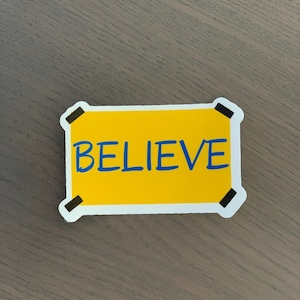 Pode incluir: Um autocolante amarelo com a palavra "BELIEVE" em azul, com letras manuscritas. O autocolante &eacute; retangular com cantos arredondados e bordas brancas. Acentos pretos em forma de fita adesiva em cada canto.