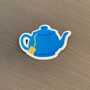 Pode incluir: Um autocolante de bule azul com uma borda branca. O bule tem uma al&ccedil;a, um bico e uma tampa. Um saquinho de ch&aacute; amarelo com as palavras "Hot Brown Water" est&aacute; preso ao bule.