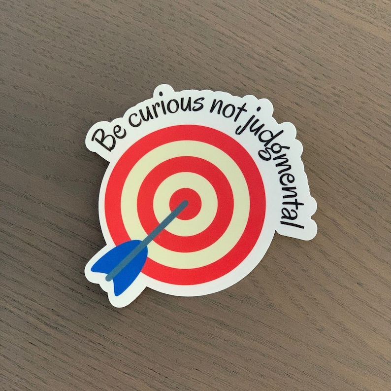 Pode incluir: Um autocolante com um alvo vermelho e creme e um dardo azul. O autocolante tem o texto "Be curious not judgmental" numa fonte manuscrita. O autocolante &eacute; branco com uma borda arredondada.