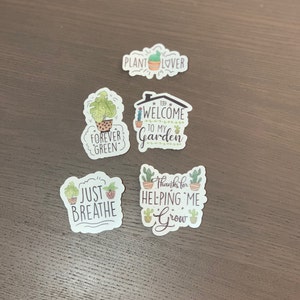 May include: Five plant-themed stickers with different sayings. The stickers are white with green and brown accents. The sayings are "Plant Lover", "Forever Green", "Welcome to My Garden", "Just Breathe", and "Thanks for Helping Me Grow".
