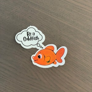 Pode incluir: Um autocolante recortado com um peixe dourado de desenho animado com escamas e barbatanas laranja, um rosto sorridente e uma nuvem de pensamento. A nuvem de pensamento diz "Be a Goldfish" em texto preto. O autocolante est&aacute; numa superf&iacute;cie de madeira.