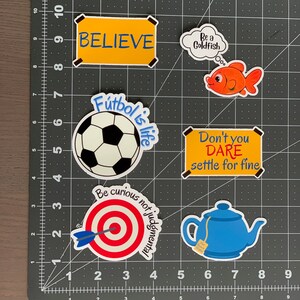 Pode incluir: Uma cole&ccedil;&atilde;o de autocolantes coloridos com v&aacute;rios desenhos e texto. Os desenhos incluem uma bola de futebol, um peixe dourado, um alvo com uma seta, uma chaleira e sinais com as palavras "BELIEVE" e "Don't you DARE settle for fine."