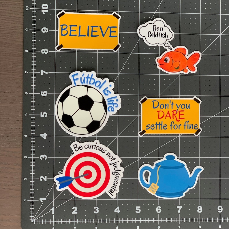 Pode incluir: Uma cole&ccedil;&atilde;o de autocolantes coloridos com v&aacute;rios desenhos e texto. Os desenhos incluem uma bola de futebol, um peixe dourado, um alvo com uma seta, uma chaleira e sinais com as palavras "BELIEVE" e "Don't you DARE settle for fine."