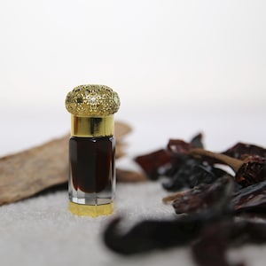 May include: A small, clear glass bottle with a gold-colored rim and a glittery gold cap. The bottle is filled with a dark brown liquid. Dried red peppers and a piece of wood are next to the bottle.