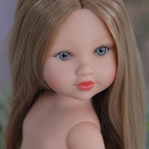 May include: A doll with long, light blonde hair and blue eyes. The doll has a fair complexion, rosy cheeks, and red lips. The doll is looking towards the viewer. The doll's hair is parted down the middle.