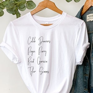 May include: White t-shirt with black script text that reads "Caleb Bowers, Paige Perry, Reid Godwin, Tyler Simmons". The t-shirt is hanging on a clothes rack with a denim jacket and a straw hat.