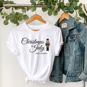 May include: White t-shirt with a black graphic that says "Christmas in July it's a thing." A nutcracker illustration is next to the text. The shirt is on a hanger with a denim jacket and a straw hat.