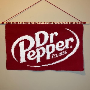 May include: A red crochet wall hanging with the Dr Pepper logo in white, suspended from a wooden dowel with red yarn. The text "Est. 1885" is also visible. A decorative item for a home.