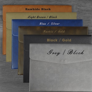 Peut inclure: Six dossiers en cuir de couleurs différentes avec des coutures contrastées. Les dossiers sont empilés les uns sur les autres. Le dossier du haut est brun avec des coutures noires et dit "Rawhide Black". Le deuxième dossier est beige avec des coutures noires et dit "Light Brown / Black". Le troisième dossier est bleu avec des coutures argentées et dit "Blue / Silver". Le quatrième dossier est vert avec des coutures dorées et dit "Rustic / Gold". Le cinquième dossier est noir avec des coutures dorées et dit "Black / Gold". Le sixième dossier est gris avec des coutures noires et dit "Grey / Black".