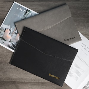May include: Two leather laptop sleeves, one grey and one black, with gold lettering. The grey sleeve has the text "Grey/Black" and the black sleeve has the text "Black/Gold".