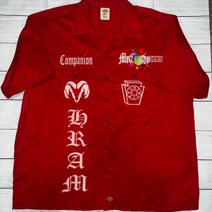 May include: Red work shirt with a pink embroidered design. The design includes a ram's head, a stylized letter "M" and the text "Companion".