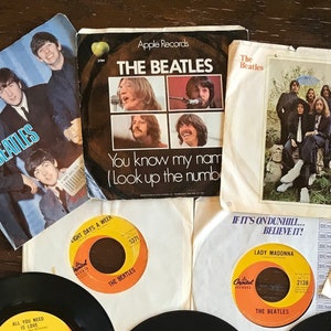 Peut inclure: Une collection de dix disques vinyles noirs avec des étiquettes jaunes et orange représentant les Beatles. Les disques sont disposés en cercle sur une surface en bois. Les étiquettes présentent des titres de chansons tels que "Love Me Do", "Paperback Writer", "Twist and Shout", "I Am The Walrus" et "Nowhere Man".