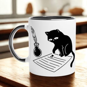 May include: White ceramic mug with a black interior and handle. The mug features a black cat illustration, ink bottle, and feather quill design. The cat is sitting on a piece of paper with paw prints. The mug is on a wooden surface.