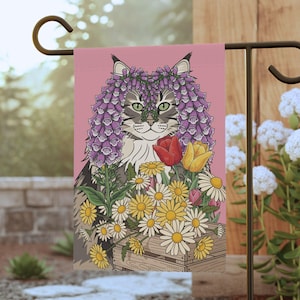 May include: A garden flag featuring a cat with a floral crown and a bouquet of flowers. The cat has gray and white fur and green eyes. The flowers include daisies, tulips, and other blooms in a wooden crate against a pink background.