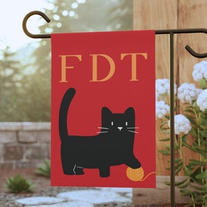 May include: A red garden flag with the letters "FDT" in gold. A black cat illustration plays with a ball of yarn. The flag is hanging on a black metal garden stake, with white flowers in the background.