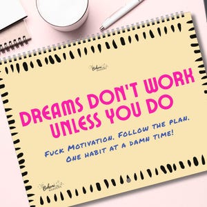 May include: A spiral-bound notepad with a cream-colored page featuring the text "DREAMS DON'T WORK UNLESS YOU DO" in pink, with additional blue text. The notepad has a black border and is on a pink surface.