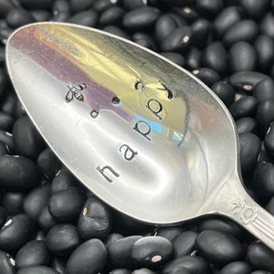 May include: A silver spoon with the word "happy" engraved on it, along with a small bee design. The spoon is resting on a bed of black beans.