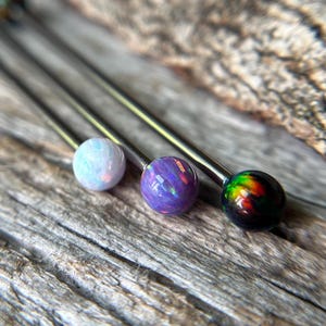 May include: Close-up of four industrial barbells with colorful opal-like ball ends. The balls are white, purple, and iridescent, set on silver-colored bars. The background is a weathered wooden surface.