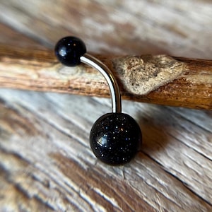 May include: A black belly button ring with a silver bar and two black round beads. The beads have a sparkly, speckled texture.