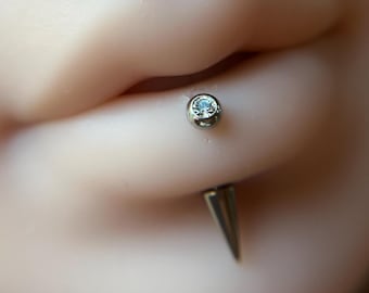 Puede incluir: Una pieza de joyería para piercing labret de tono plateado insertada en un modelo de silicona de un labio inferior. La joya presenta una pequeña piedra preciosa redonda y transparente en una base metálica circular y una punta cónica afilada.