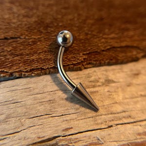 Puede incluir: Un piercing corporal de plata con una barra curva y una bola redonda en un extremo y un extremo puntiagudo en el otro.