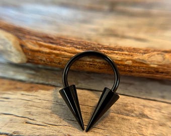 May include: Black circular barbell with two pointed ends. The barbell is made of surgical steel and has a black finish.
