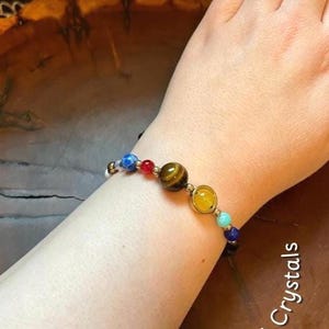 May include: A bracelet with beads representing the planets in the solar system. The beads are made of different colors and materials, including blue, red, yellow, and brown. The bracelet is on a person's wrist. © Cali Crystals