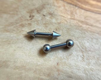 Puede incluir: Dos piezas de joyería corporal tipo barra de metal plateado. Una pieza presenta dos extremos en forma de cono puntiagudo, mientras que la otra pieza presenta dos extremos esféricos lisos y redondeados. Ambas tienen una barra central.
